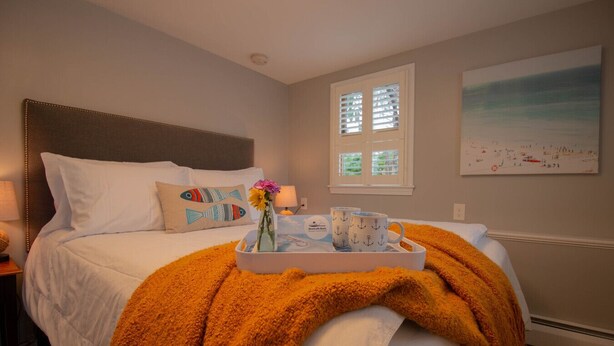 Ein Schlafzimmer mit einem Bett, Kissen, einem Tablett mit Tassen, einer Vase mit Blumen und einem Fenster mit Jalousien.