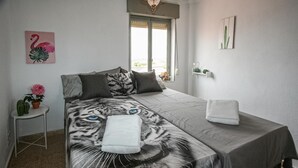 A bedroom with a bed featuring a tiger's face design, a small bedside table with a potted plant, a window with curtains, and a wall-mounted shelf with decorative items. - Cozy apartment in Santa Pola with WiFi (Santa Pola)