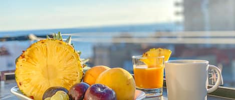 Mesa de café da manhã com uma tigela de frutas, um copo de suco de laranja e uma caneca em uma varanda com vista para o mar.