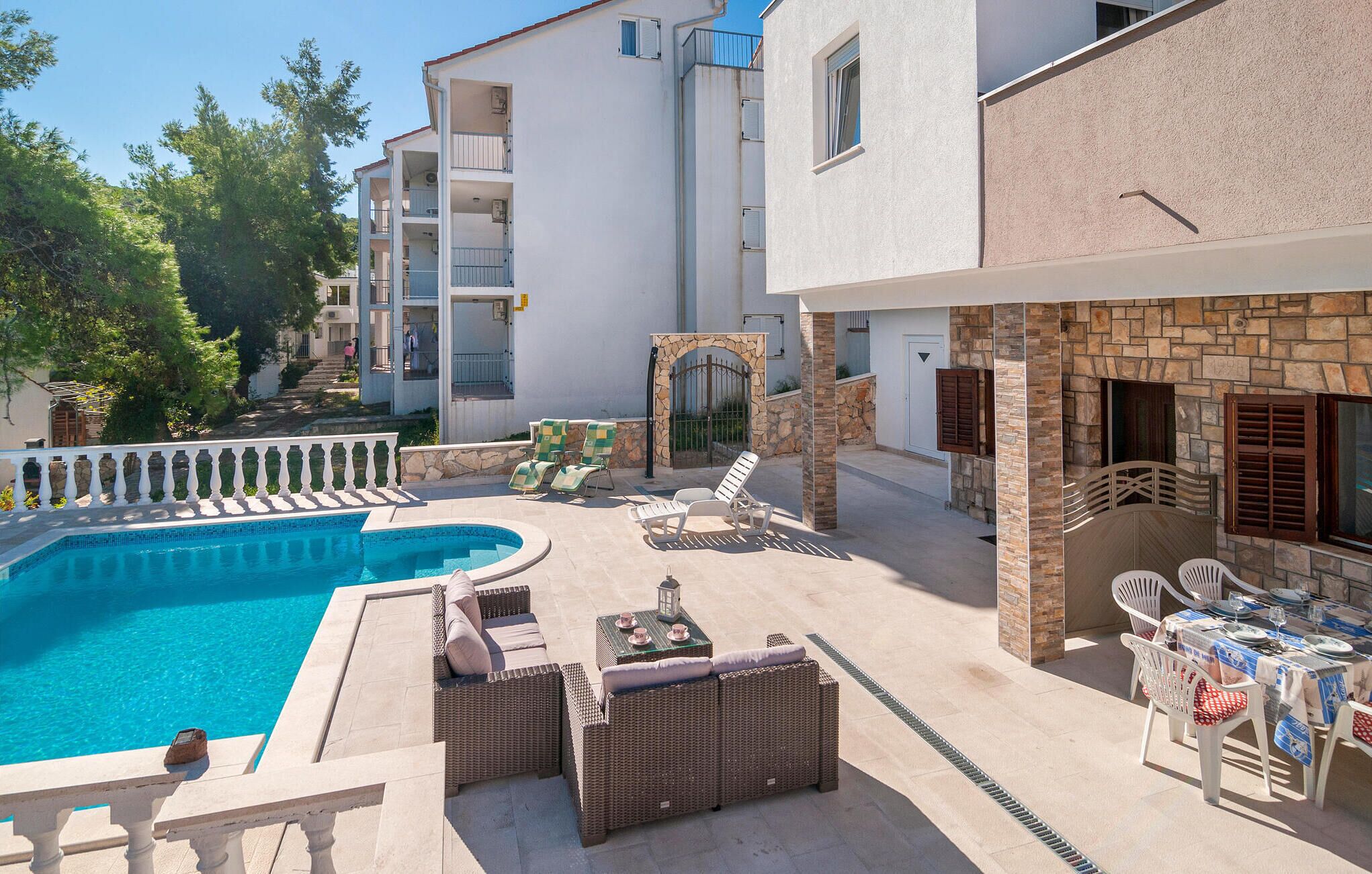 Area piscina con lettini e un tavolo, circondata da edifici con balconi e un muro in pietra.