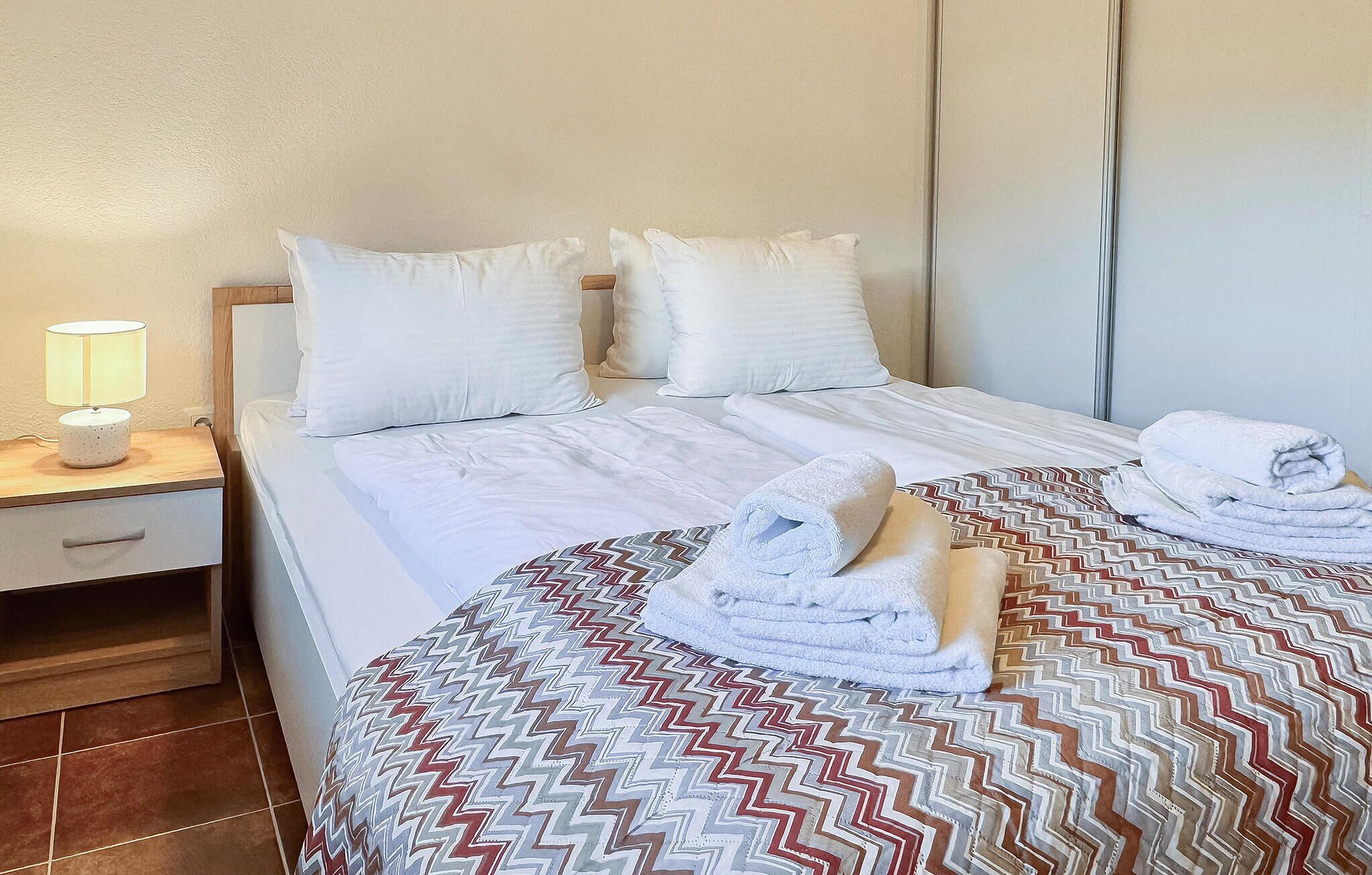 Cama bem arrumada com lençóis brancos e duas toalhas enroladas, um abajur e um edredom estampado.