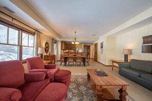 Uma sala de estar espaçosa com uma mesa de jantar de madeira, poltronas vermelhas e um sofá verde. Uma cozinha é visível ao fundo, com armários de madeira e um candelabro.