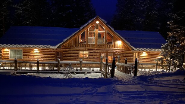 A log cabin with a snow-covered roof, wooden exterior, and a fenced front yard. - Rustic Mountain Cabin (New Meadows)