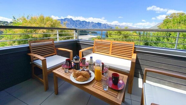 A balcony with wooden furniture, a breakfast setup including croissants, coffee, and juice, and a scenic mountain view.