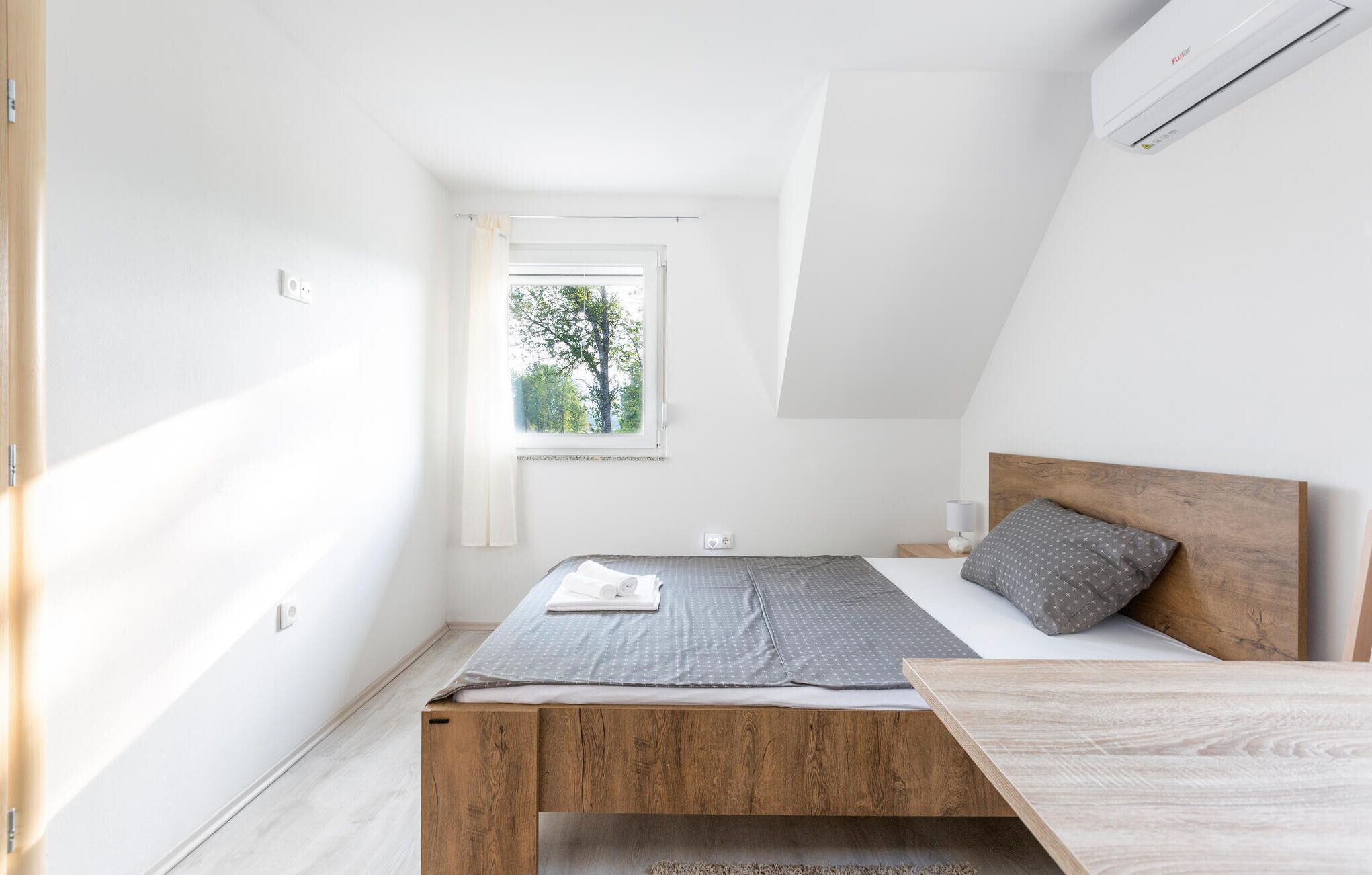 A small, modern bedroom with a sloped ceiling, a single bed, a nightstand, and a window with a view of trees.