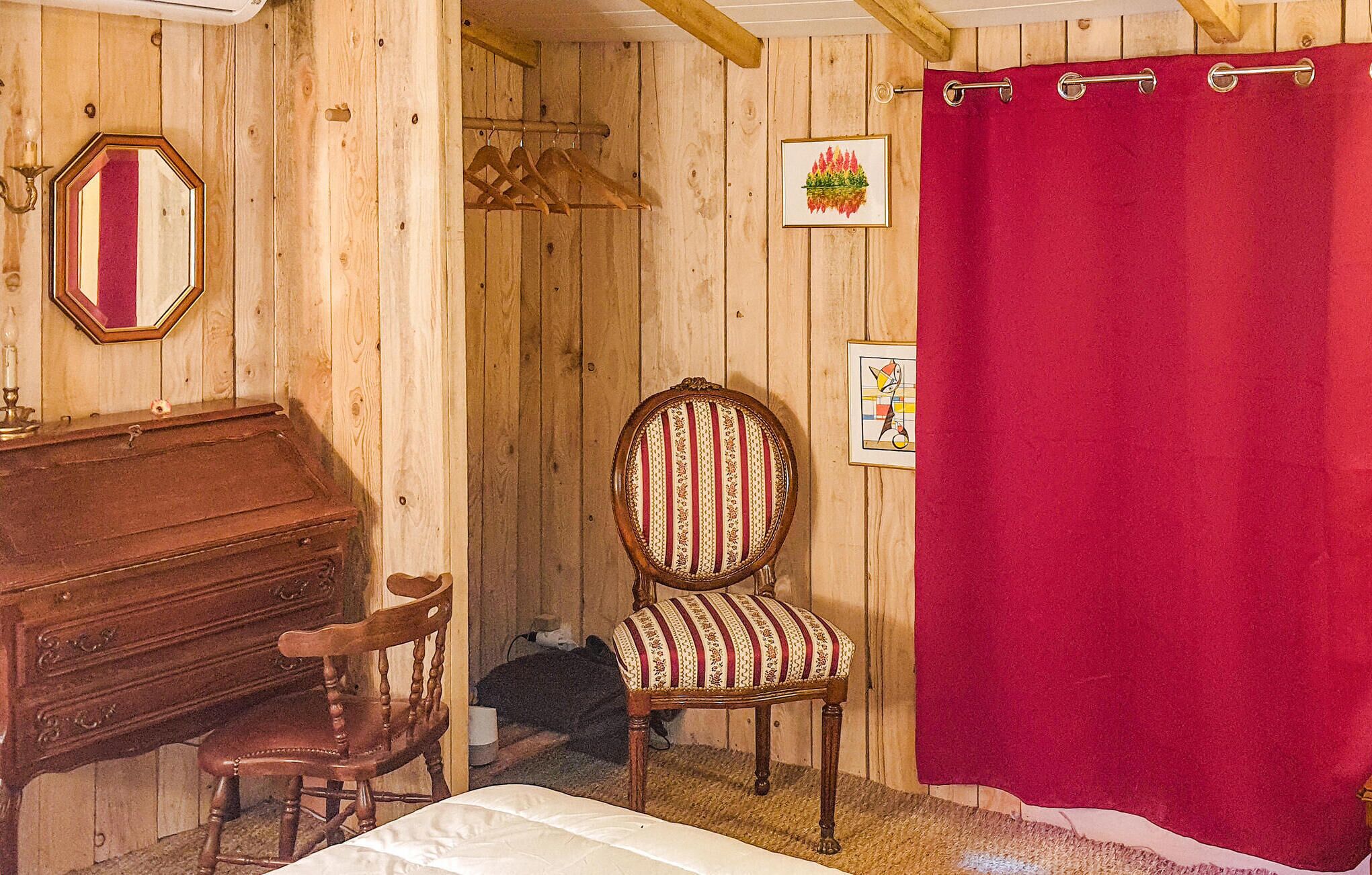 A wooden room with a bed, a wooden chair, a wooden desk, a mirror, and a red curtain.