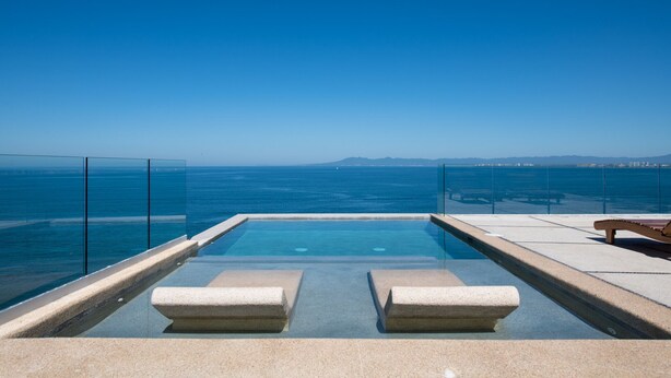 An infinity pool with clear blue water, a glass railing, and a view of the ocean and distant mountains.
