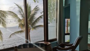 A room with a large window overlooking a beach with palm trees. There is a chair and a small table in the foreground.