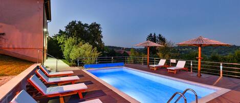 A swimming pool with lounge chairs and umbrellas, surrounded by a wooden deck and overlooking a scenic landscape.