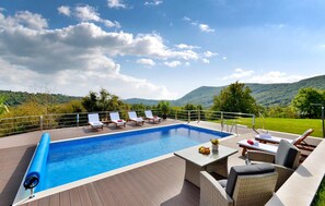 A swimming pool with lounge chairs and a table, surrounded by a wooden deck and a metal railing.