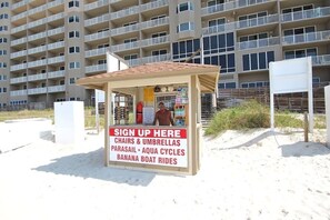 A beachside kiosk with a sign offering chairs, umbrellas, parasailing, aqua cycles, and banana boat rides.