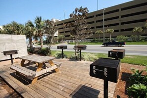 An outdoor patio with a wooden table and benches, surrounded by greenery and a parking garage in the background.