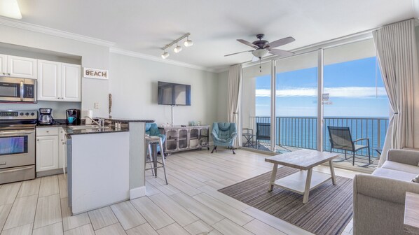 A modern kitchen with a central island, a dining area with a table and chairs, a balcony with a view of the ocean, and a ceiling fan.