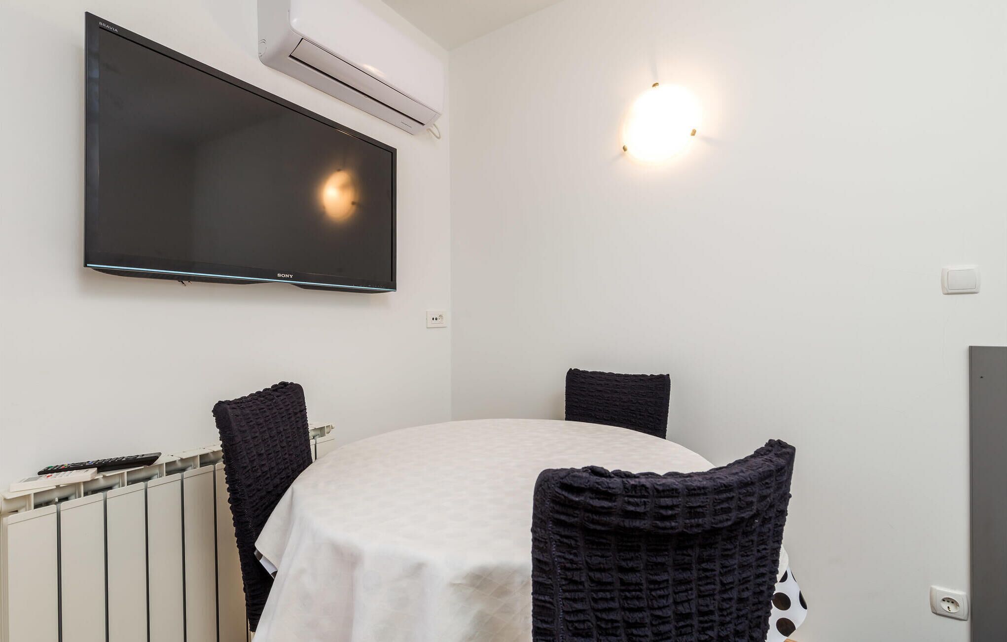 A room with a flat-screen TV mounted on the wall, a table with a white tablecloth, and two dark chairs.