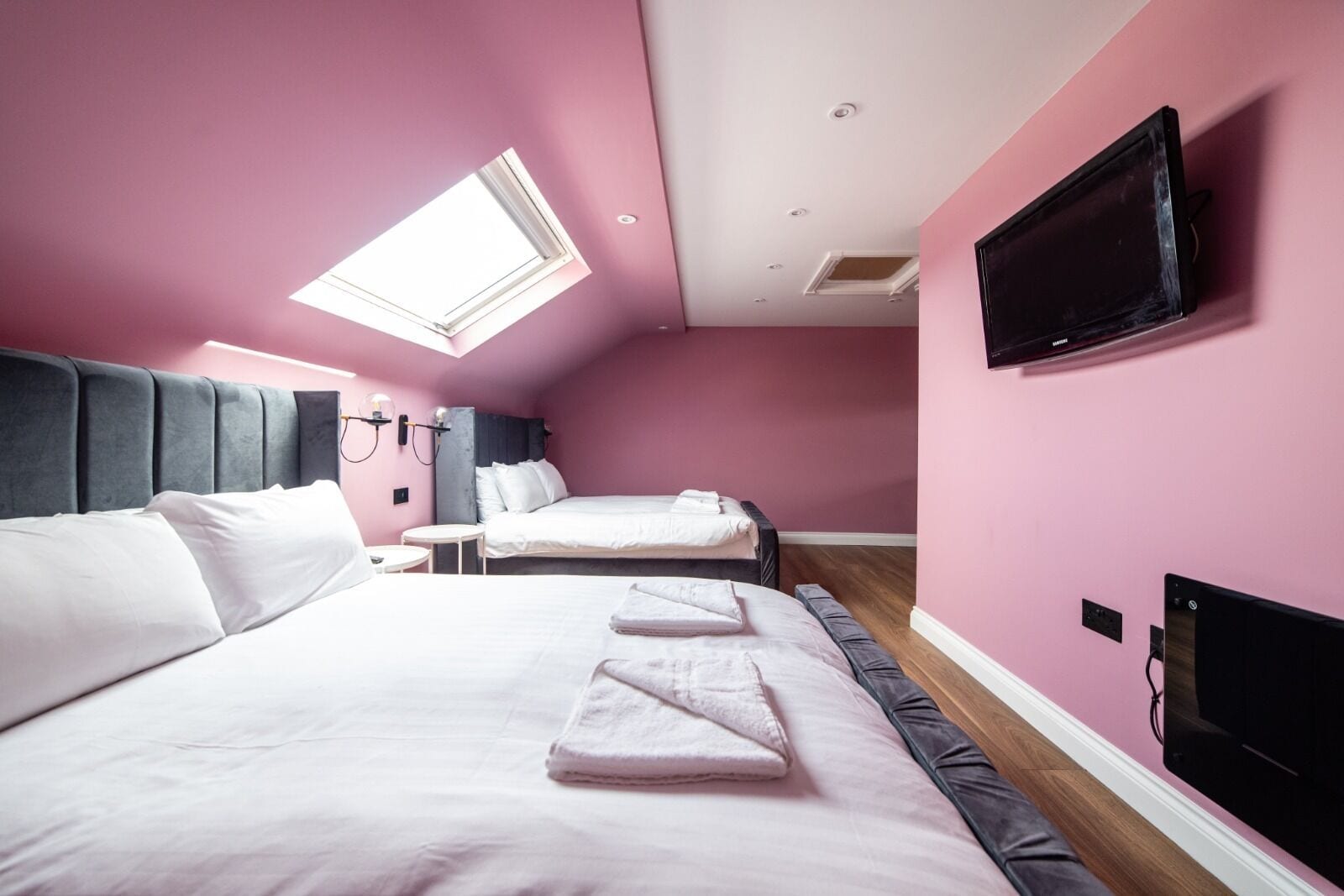 A hotel room with a large bed, a smaller bed, a desk, a chair, a flat-screen TV, and a skylight.