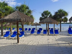 Een strand met blauwe ligbedden en rieten parasols, een zandstrand en een rustig wateroppervlak op de achtergrond.