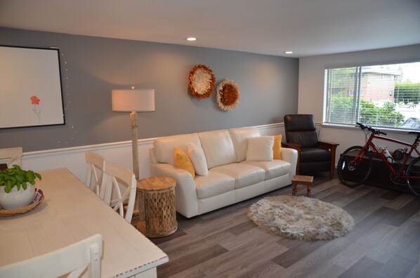 A living room with a white sofa, a dining table with chairs, a lamp, a bicycle, and a window with blinds.