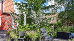 A wooden deck with two chairs and a small table, surrounded by lush greenery and a red building in the background. - Gorgeous home in Rörvik with WiFi (Rörvik)