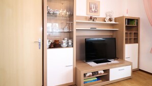 Una habitación con una televisión sobre un soporte de madera, un aparador de vidrio con vajilla, y un estante con libros y una vela.