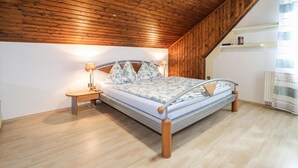 Un dormitorio con techo de madera, una cama con ropa de cama floral, una mesita de noche con lámpara y un radiador.