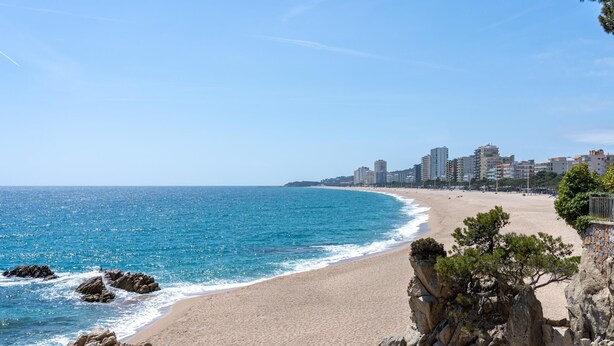 Ein Strand mit klarem, blauem Wasser, Felsenformationen und einer Stadtlandschaft im Hintergrund.