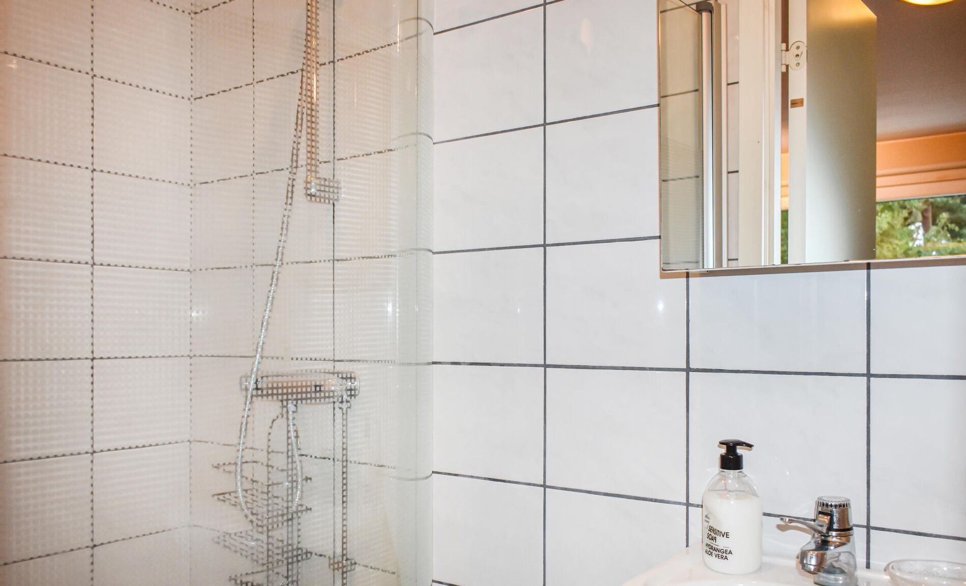 Une salle de bain moderne avec des murs recouverts de carreaux blancs, une douche en verre, une petite étagère en verre, un lavabo avec un robinet et un miroir.