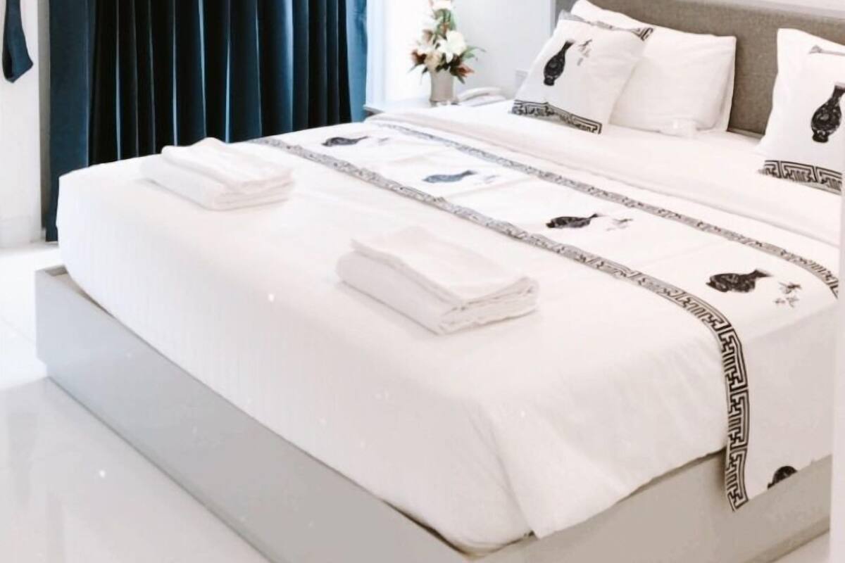Un dormitorio moderno con una cama grande, cortinas oscuras, una ventana con persianas, una mesita con un jarrón y un cuadro en la pared.