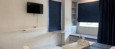 A hotel room with two beds, a TV mounted on the wall, a window with dark curtains, and a small shelf unit.