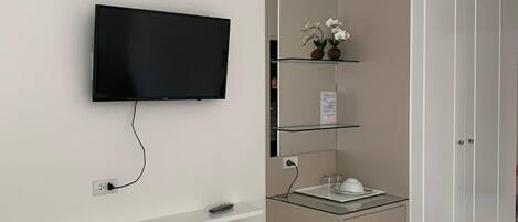 Ein modernes Hotelzimmer mit einem flachbildfernseher an der Wand, einem kleinen Regal darunter, einem Spiegel, einem Schrank mit Waschbecken und einem Mülleimer.