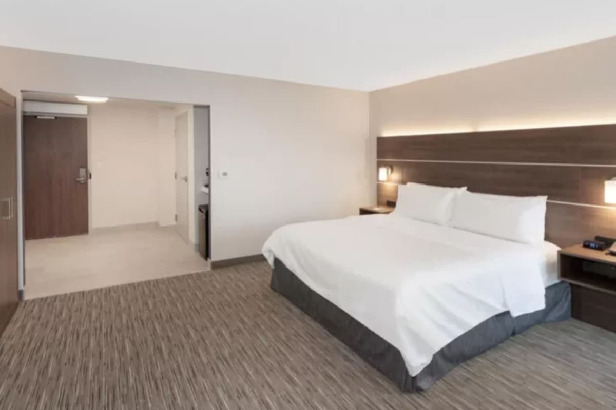 A modern hotel room with a large bed, a wooden headboard, a bedside table with a lamp, and a carpeted floor.