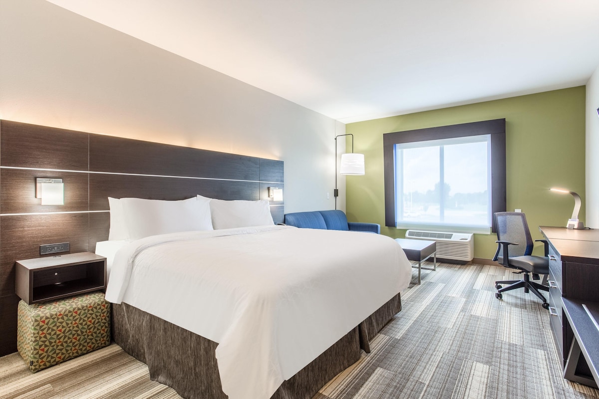 A hotel room with a large bed, a desk with a chair, a small table, a lamp, and a window with a view.