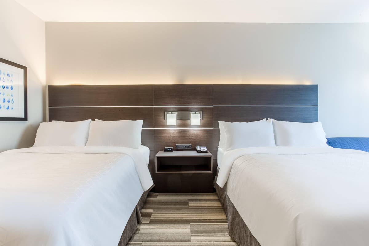 Habitación de hotel con dos camas, una mesita de noche con teléfono y una pequeña mesa, y cabecera con iluminación integrada.