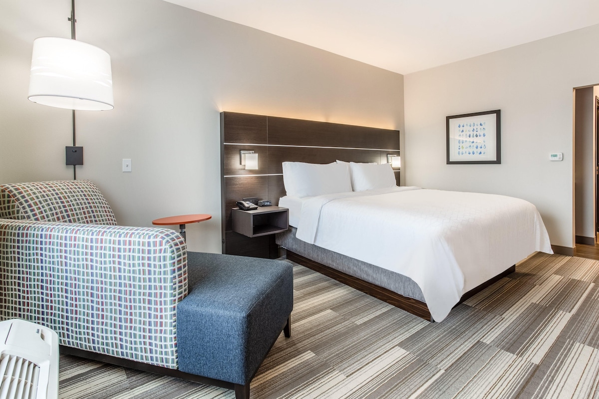 A hotel room with a large bed, a desk with a lamp, a chair, and a small table.