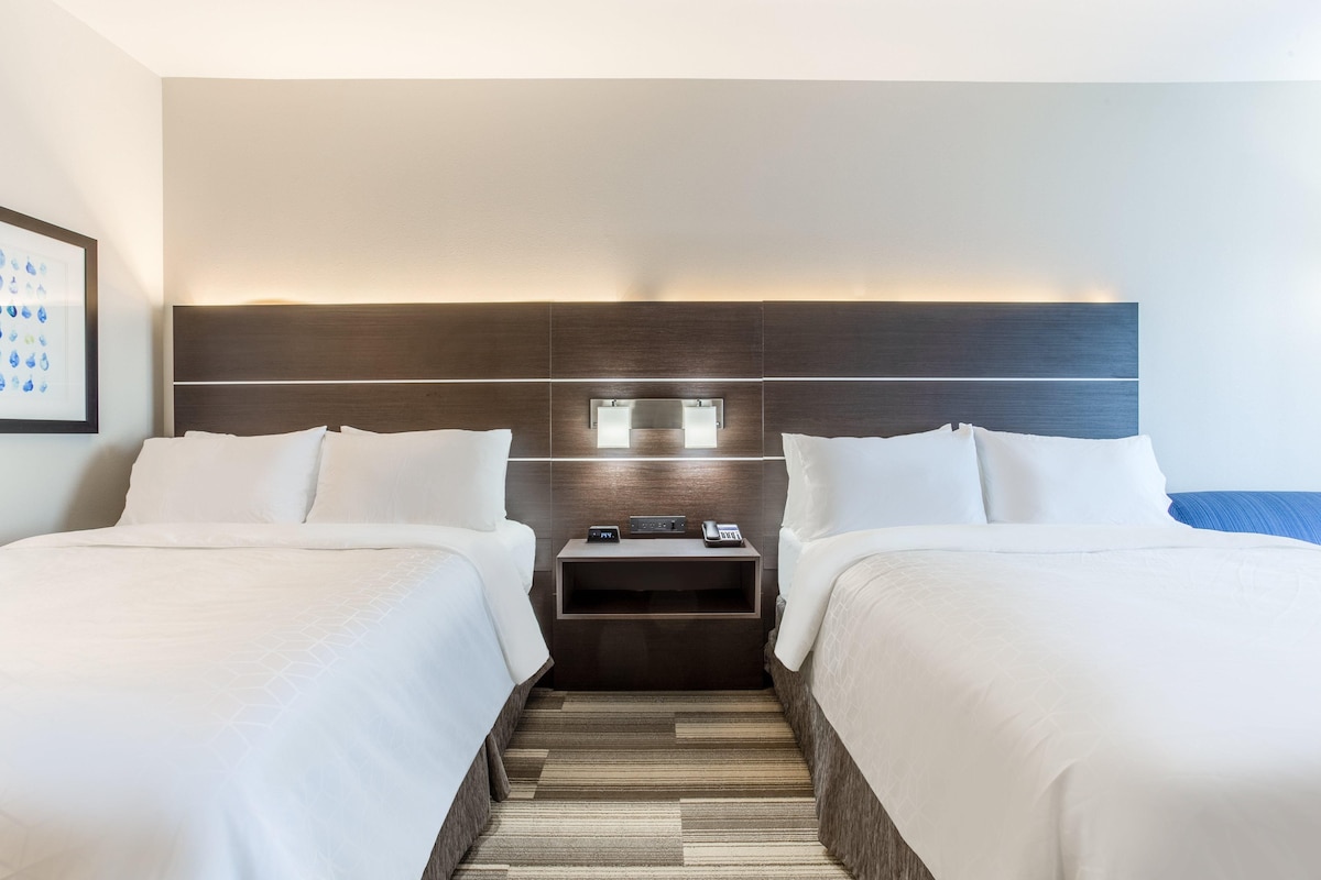 A hotel room with two beds, a nightstand with a phone and a small table, and a headboard with built-in lighting.