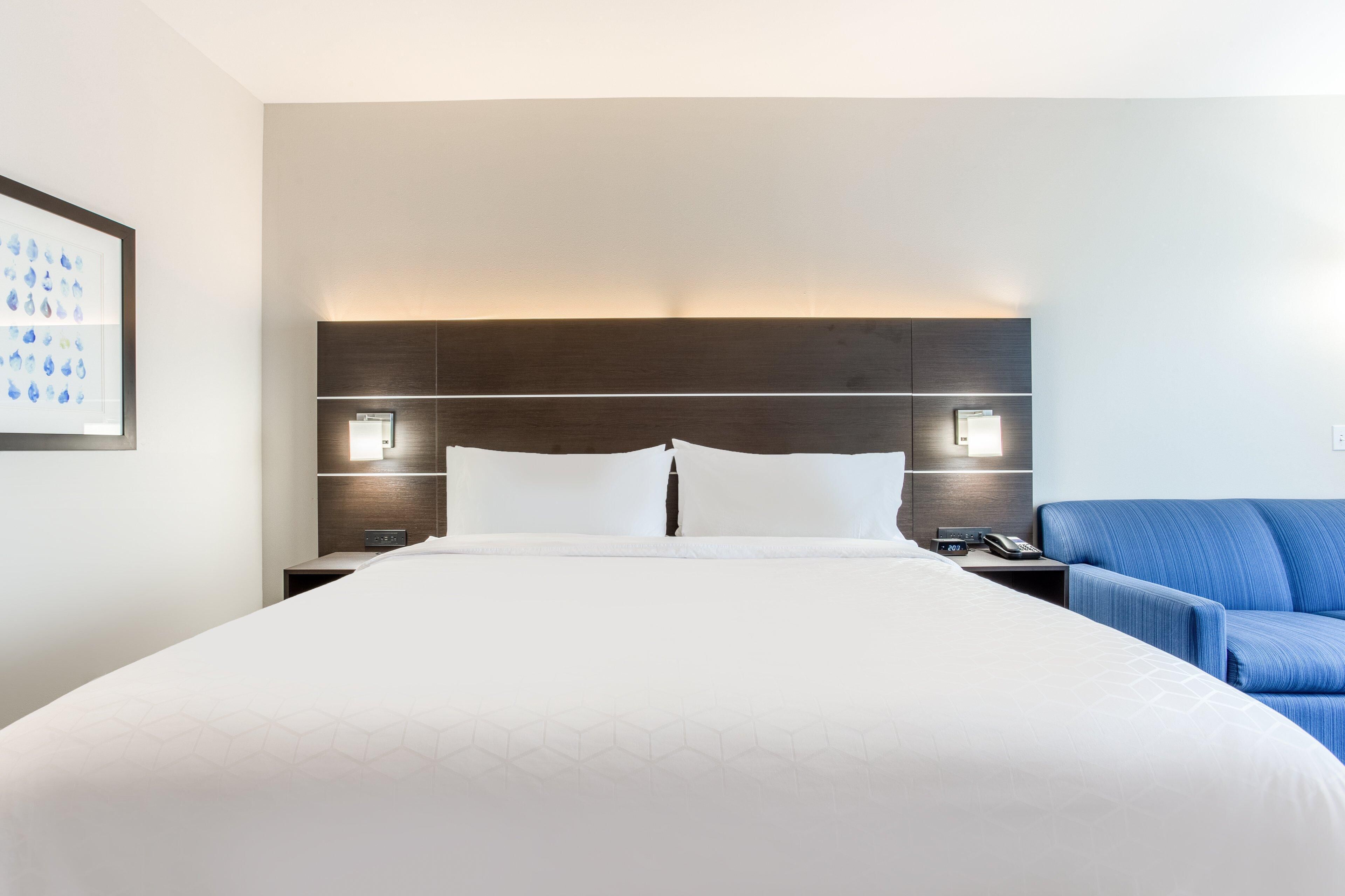 A hotel room with a large bed, two bedside tables, a nightstand, a blue armchair, and a framed picture on the wall.