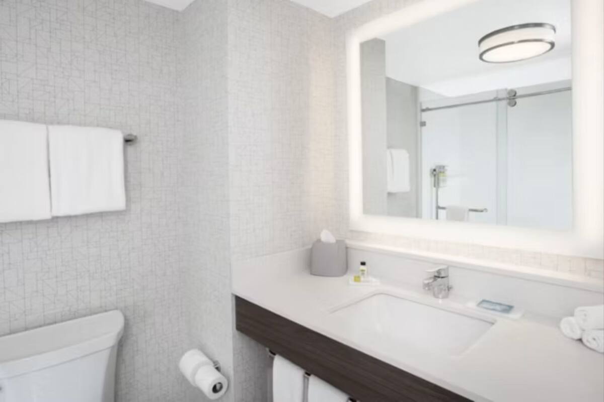 Un baño moderno con un espejo grande, baldosas blancas, un tocador de madera oscura y un inodoro.