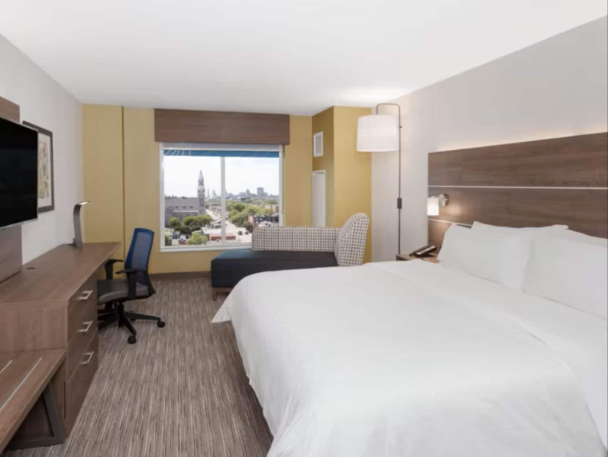 A hotel room with a large bed, a desk with a chair, a television, and a window with a city view.