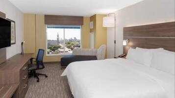 A hotel room with a large bed, a desk with a chair, a television, and a window with a city view.