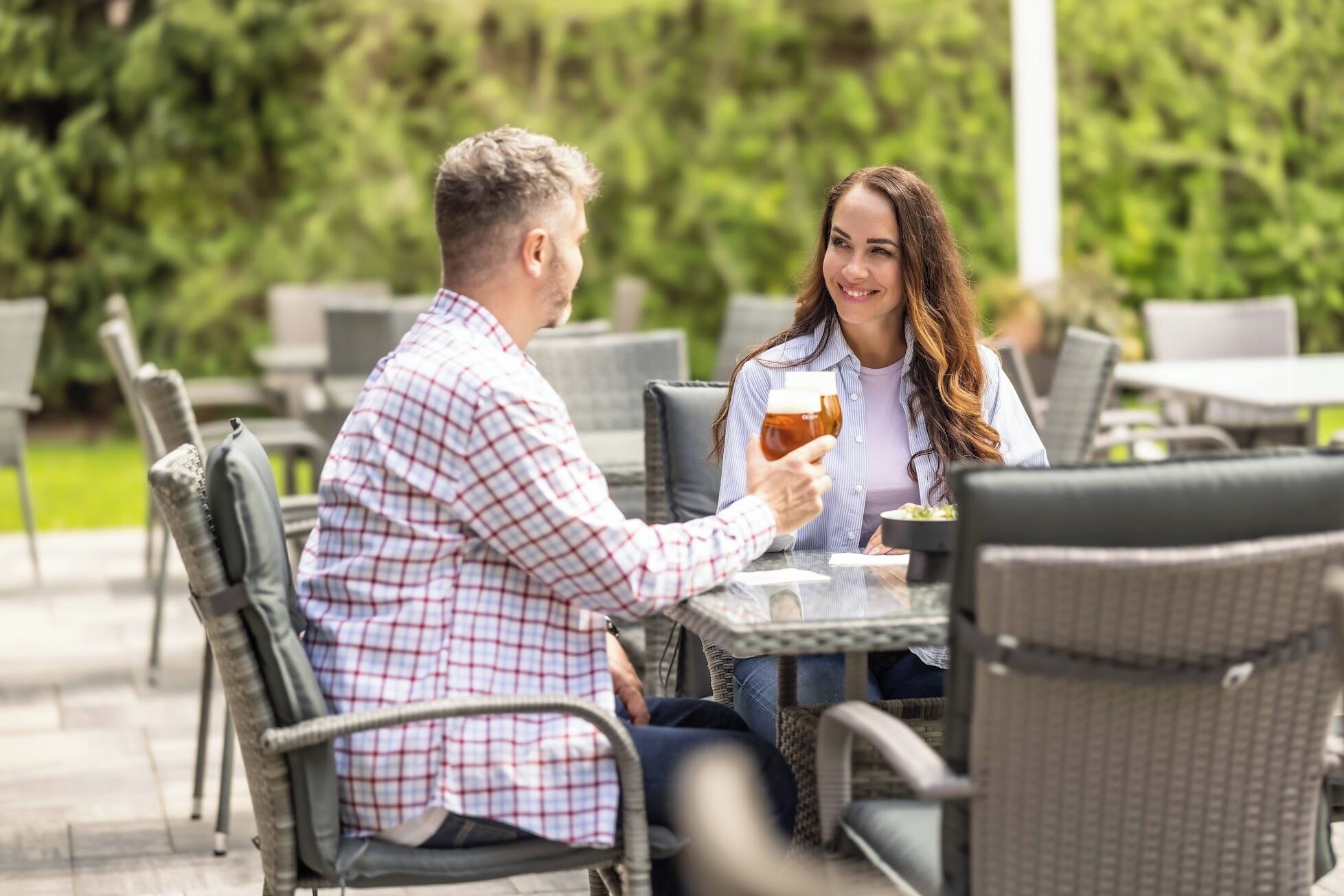 A couple seated at an outdoor cafe with wicker chairs and a table, holding a glass of beer.