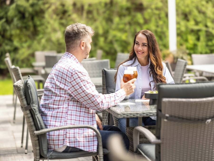 Una pareja sentada en un café al aire libre, con sillas de mimbre y una mesa, sosteniendo una jarra de cerveza.