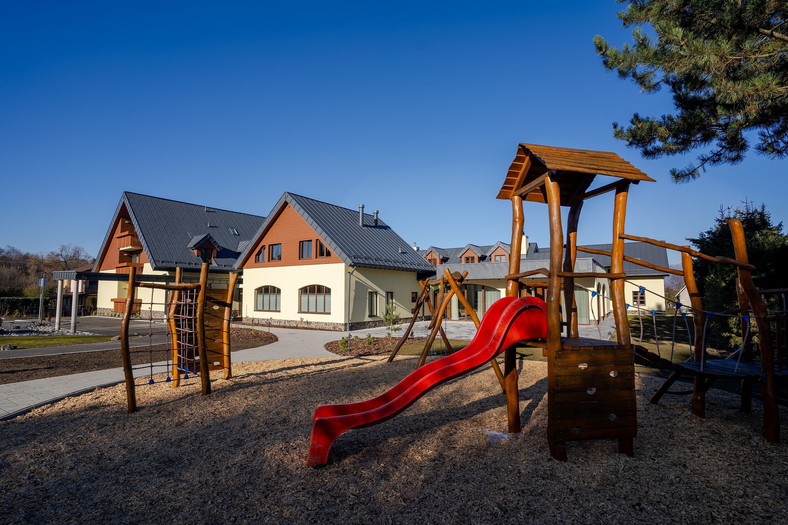 A playground with a red slide and climbing structures, set against a backdrop of a house with a sloped roof and a clear blue sky.