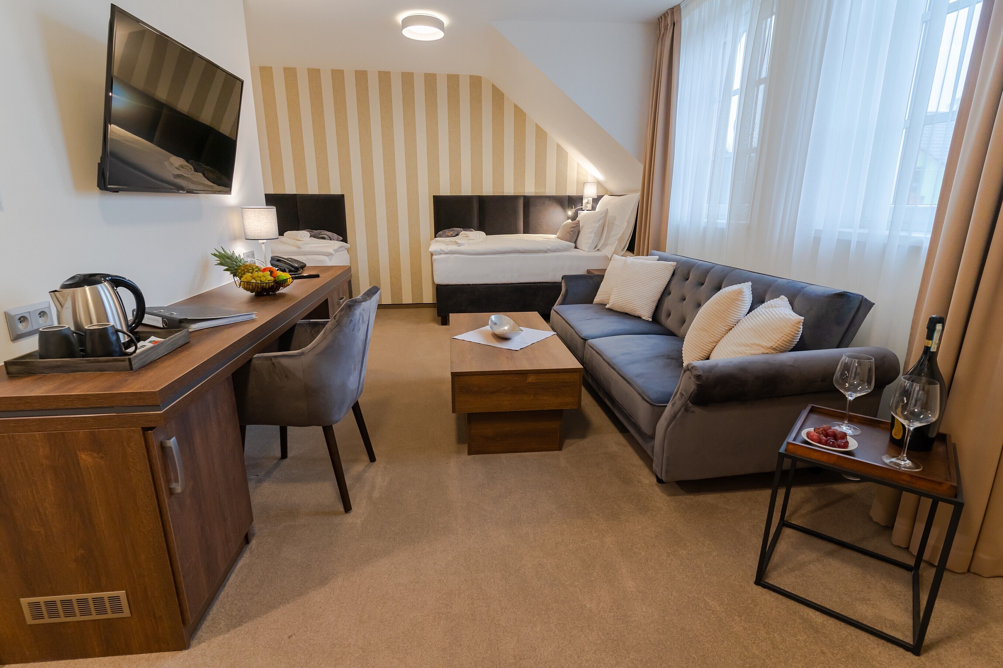 A hotel room with a bed, a sofa, a desk with a kettle, a TV, and a small table with wine glasses and fruit.