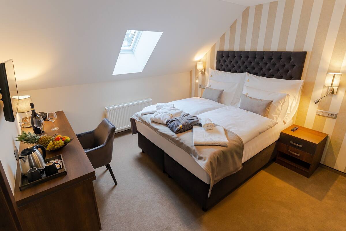 Une chambre d’hôtel moderne avec un grand lit, un bureau doté d’une machine à café, un panier de fruits et une vue sur le ciel grâce à une lucarne.