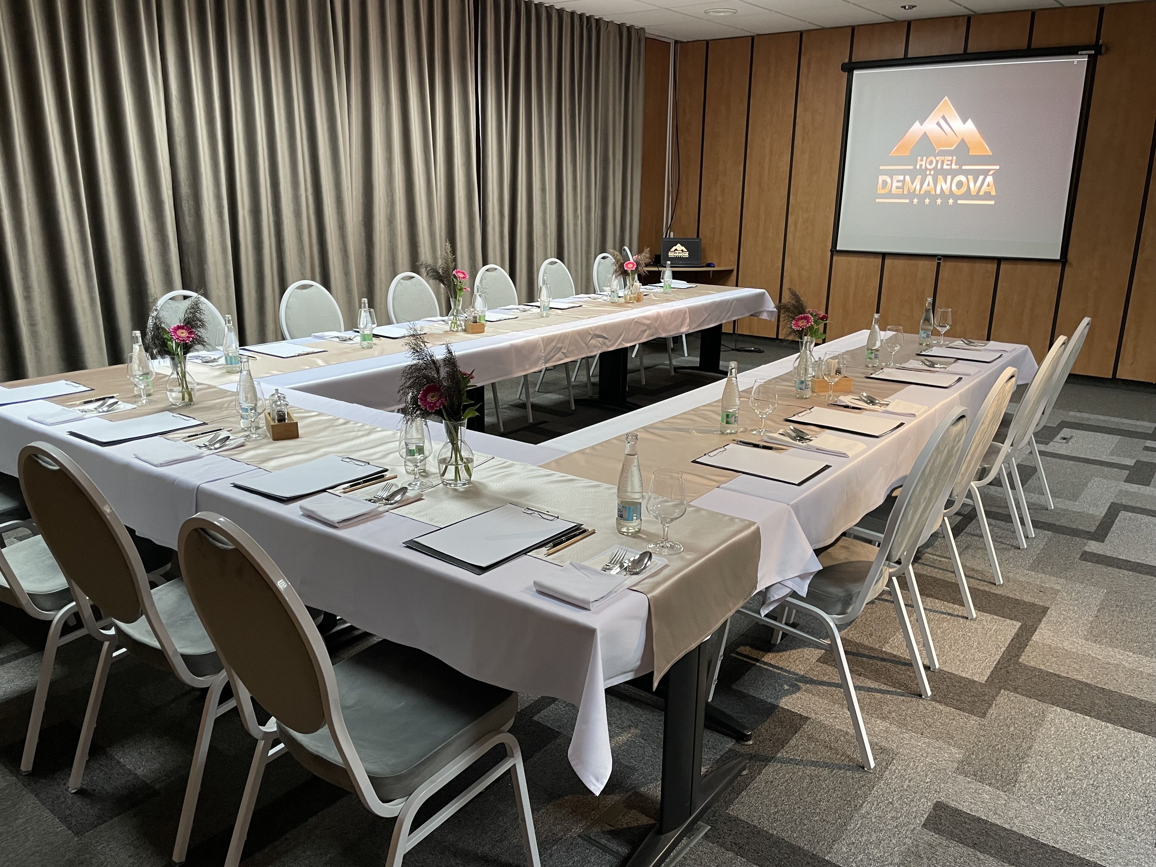 A conference room with a long table set for a meeting, white chairs, and a projector screen displaying the hotel name.