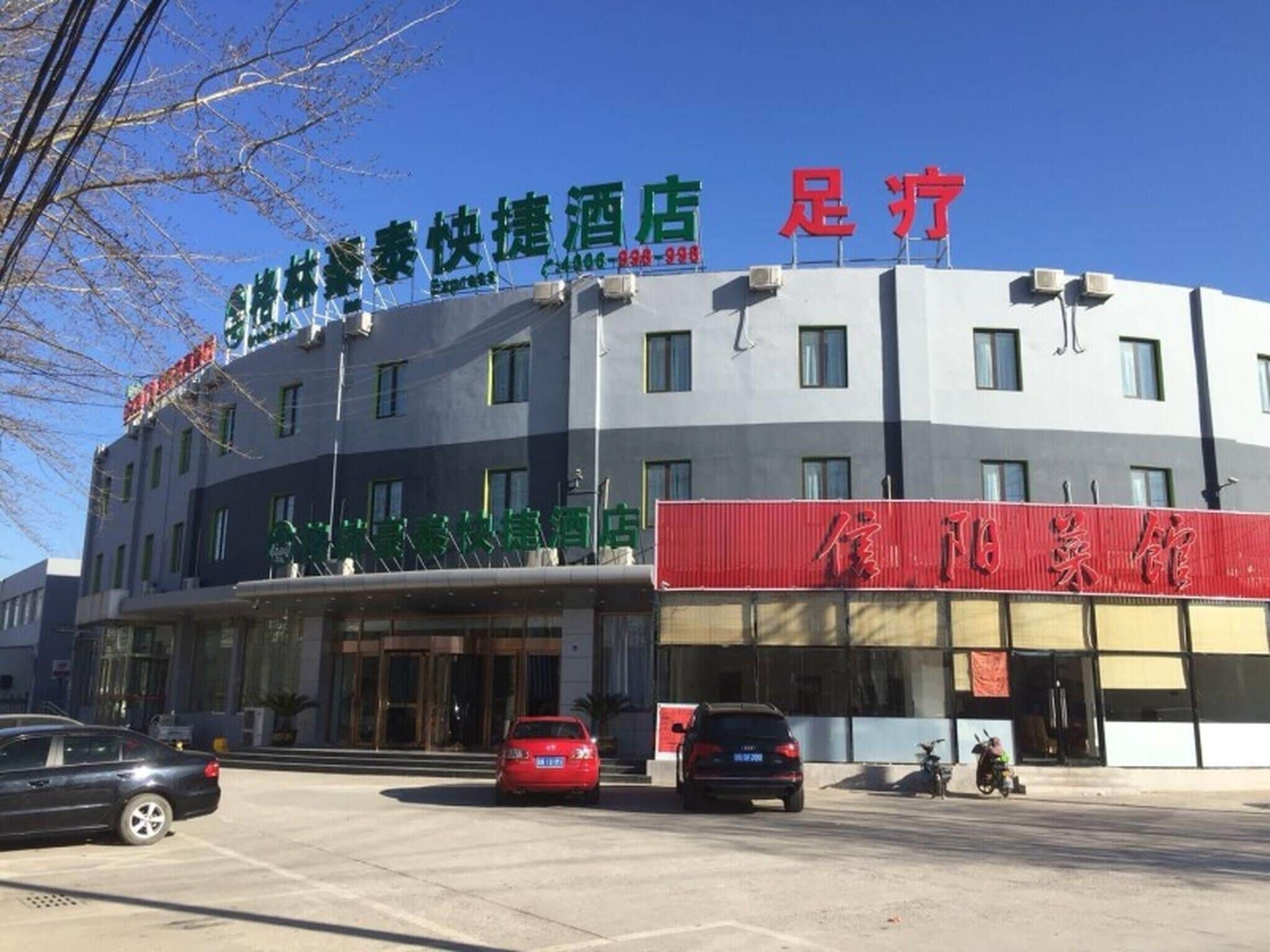 A hotel with a signboard displaying Chinese characters, a red signboard with more Chinese characters, and a few parked cars in front.