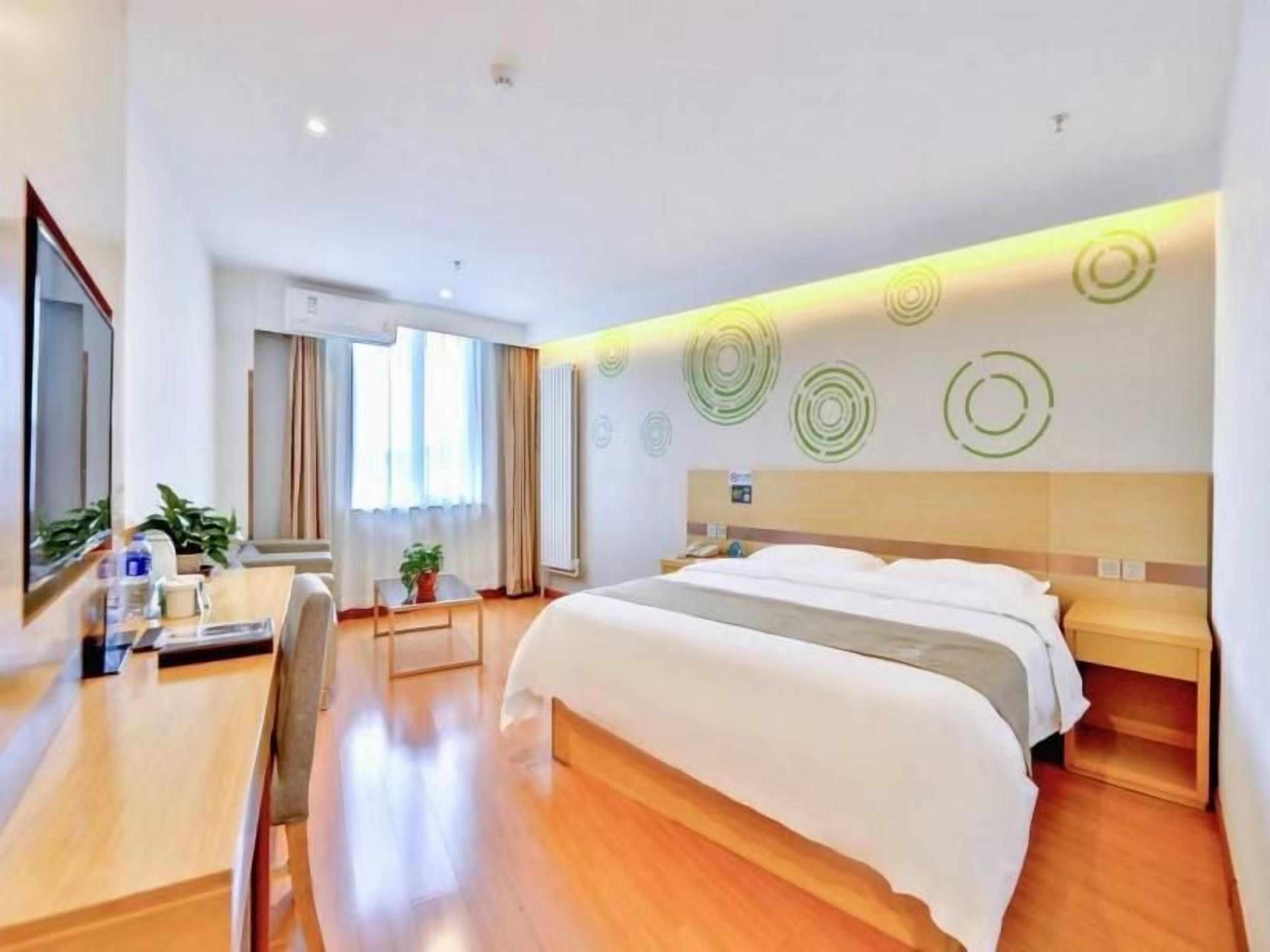 A hotel room with a large bed, a desk, and a chair. There is a window with curtains, a radiator, and wall art.