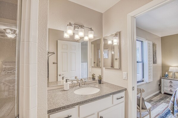 A bathroom with a white sink, vanity, and a large mirror with lights. There is a window with blinds and a chair in the corner. - Saguaro Escape | Pool/Tennis/Hiking/Patio (Tucson)