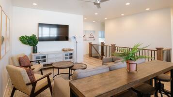 A spacious living room with a wooden dining table, comfortable seating, a TV mounted on the wall, and a staircase leading to an upper level.