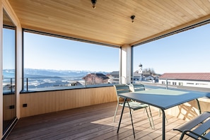 A balcony with wooden flooring, a table, and chairs, overlooking a cityscape with mountains in the distance. - Apartment 2 - Apartments Poldi (Sulzberg)