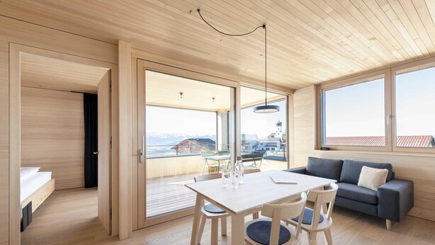 Een modern interieur met een houten vloer, een eettafel met stoelen, een bank en grote ramen met uitzicht op een berg en een gebouw.